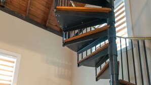 Una escalera de caracol con escalones de madera y barandillas de metal negro en una habitación con vigas de madera en el techo y una pared blanca.