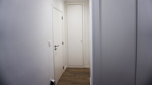 Um corredor estreito com paredes brancas e chão de madeira, que leva a uma porta fechada no final.