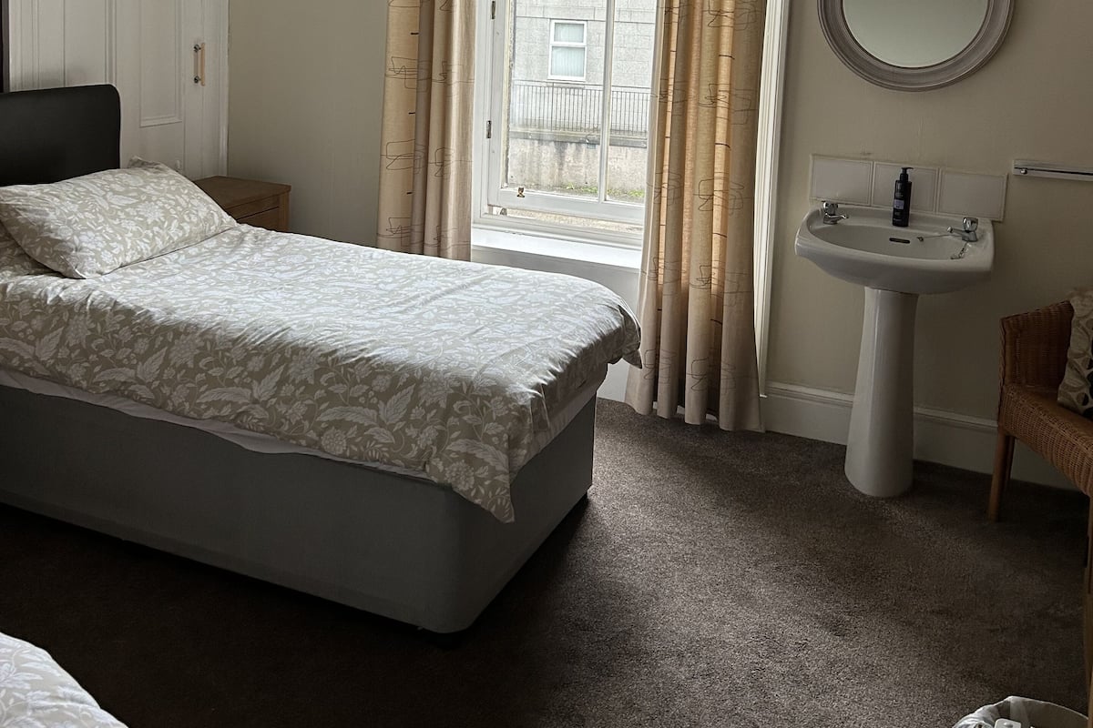 A bedroom with a bed, a bedside table, a window with curtains, a round mirror, and a sink.
