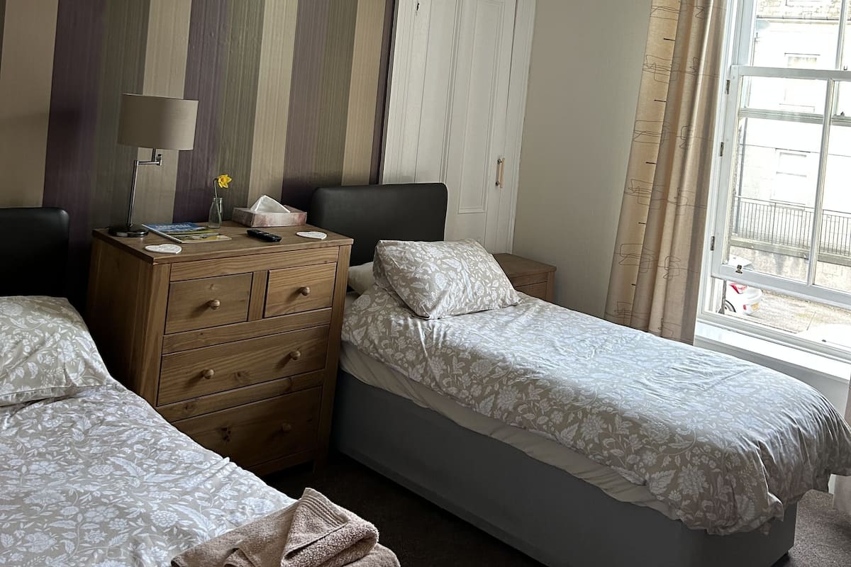 A hotel room with two single beds, a wooden dresser, a desk with a lamp, and a window with curtains.