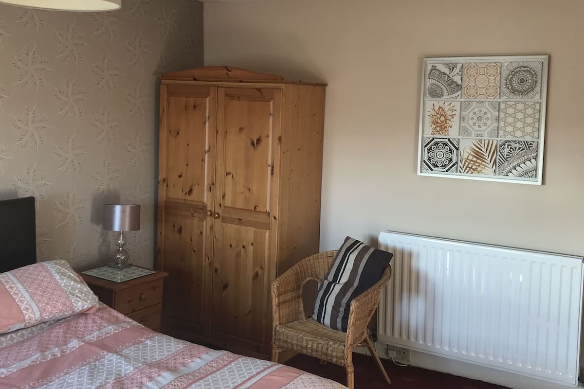 A bedroom with a bed, a wooden wardrobe, a wicker chair, a bedside table with a lamp, and a wall with a decorative plate.