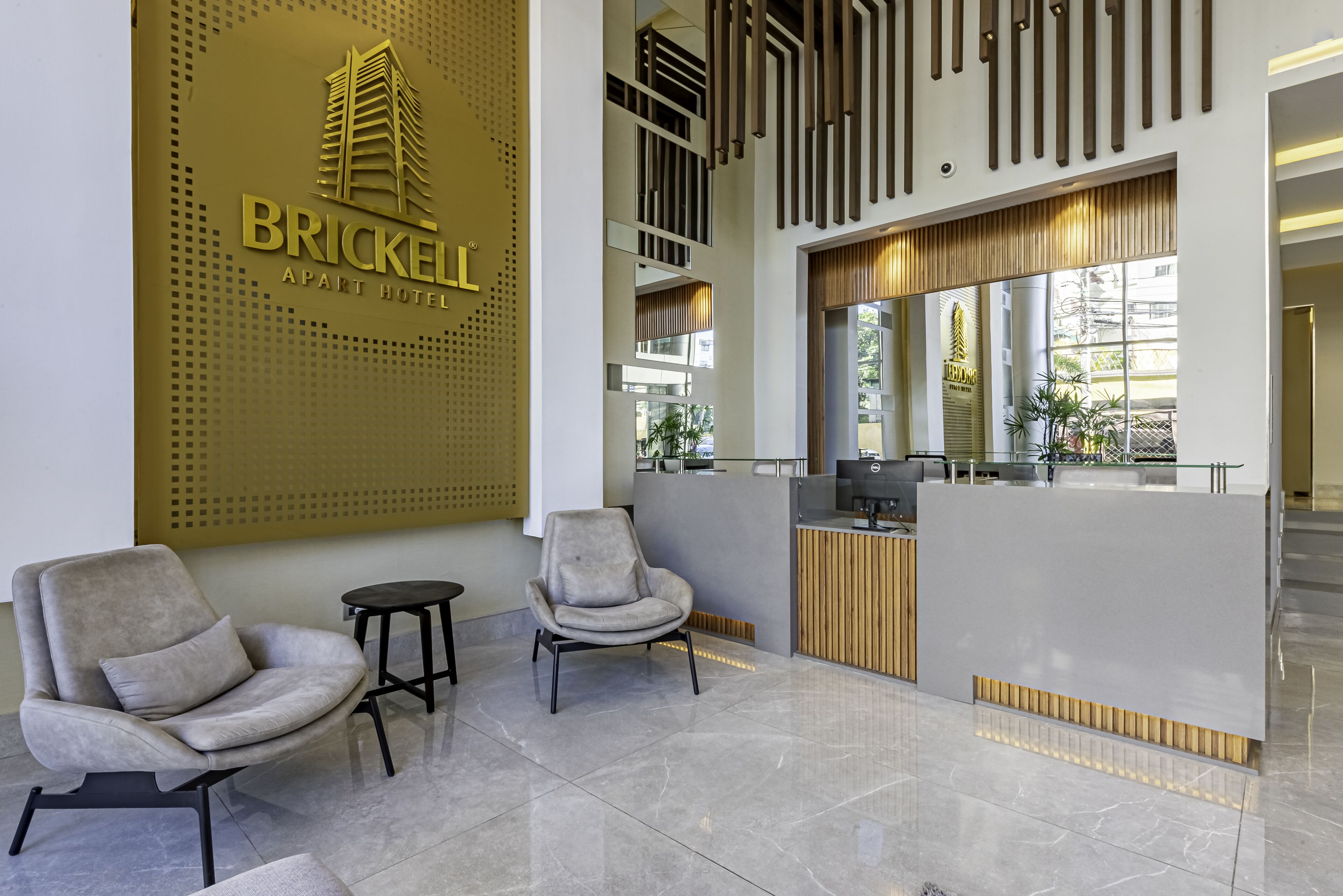A modern hotel reception area with a prominent logo, two armchairs, a reception desk, and a large window providing natural light.
