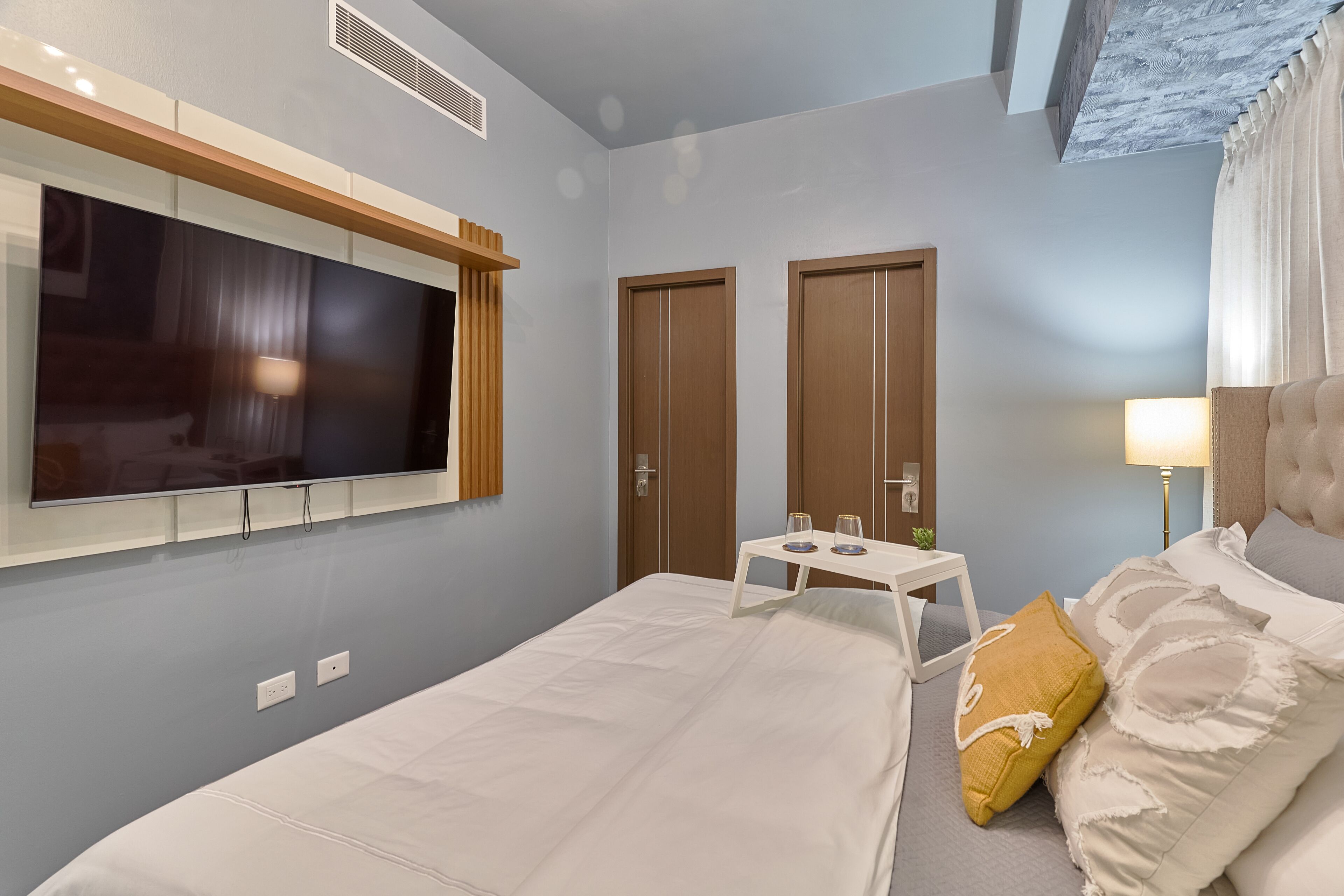 A hotel room with a large bed, a small table with two glasses, a TV mounted on the wall, and two doors.