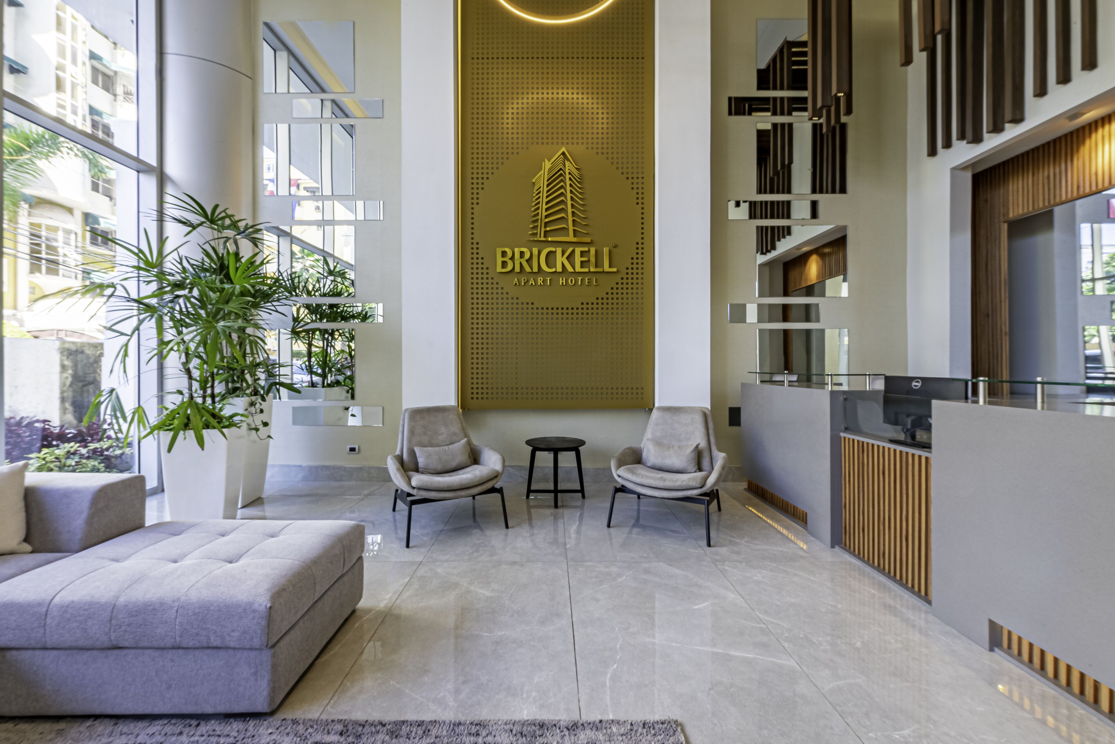 Una hall moderna con un grande logo di Brickell, un’area salotto con due poltrone e un tavolino, e un’ampia finestra con piante.