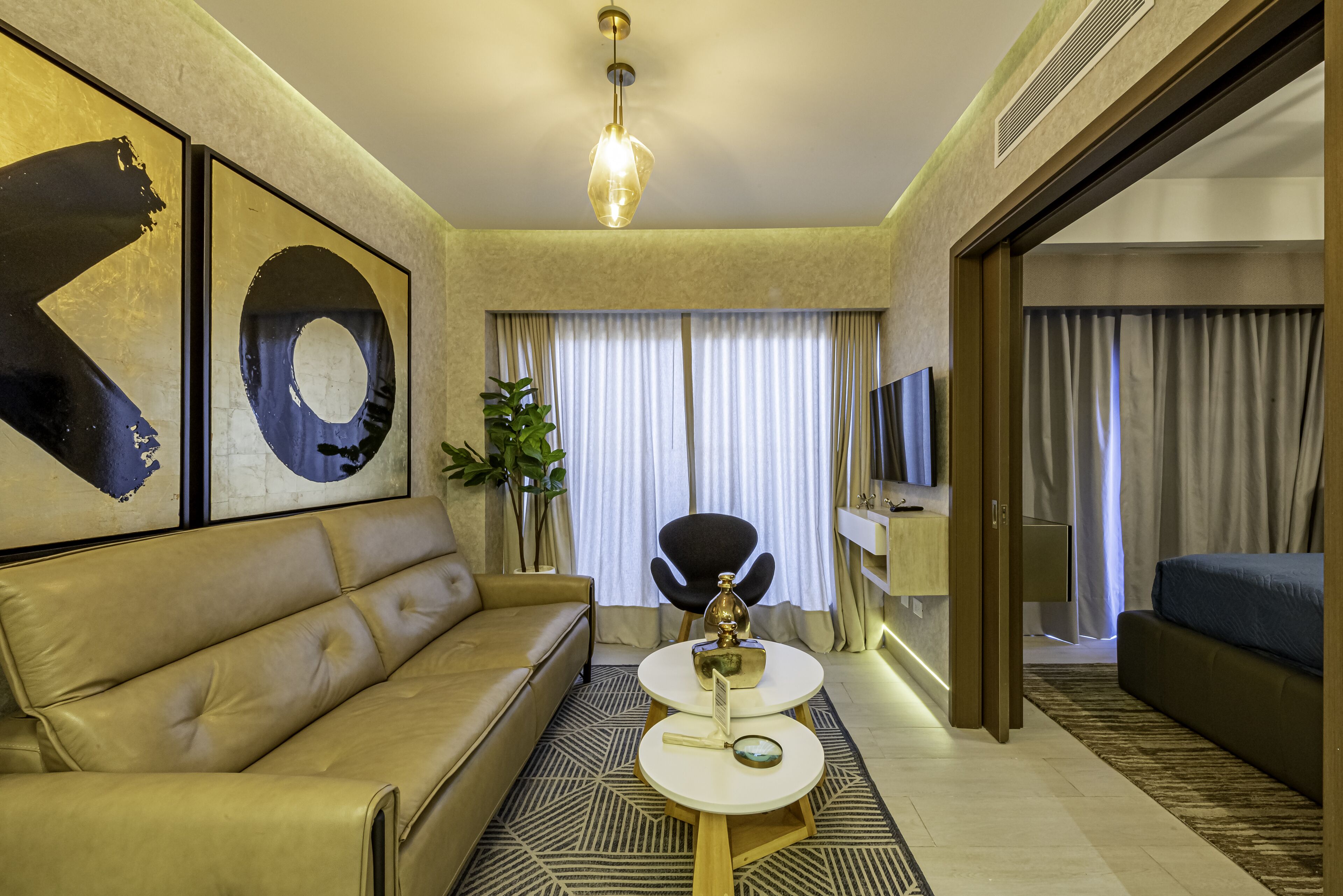 Una habitación de hotel moderna con un sofá de cuero, una mesa de centro redonda, un televisor y un ventanal con cortinas.