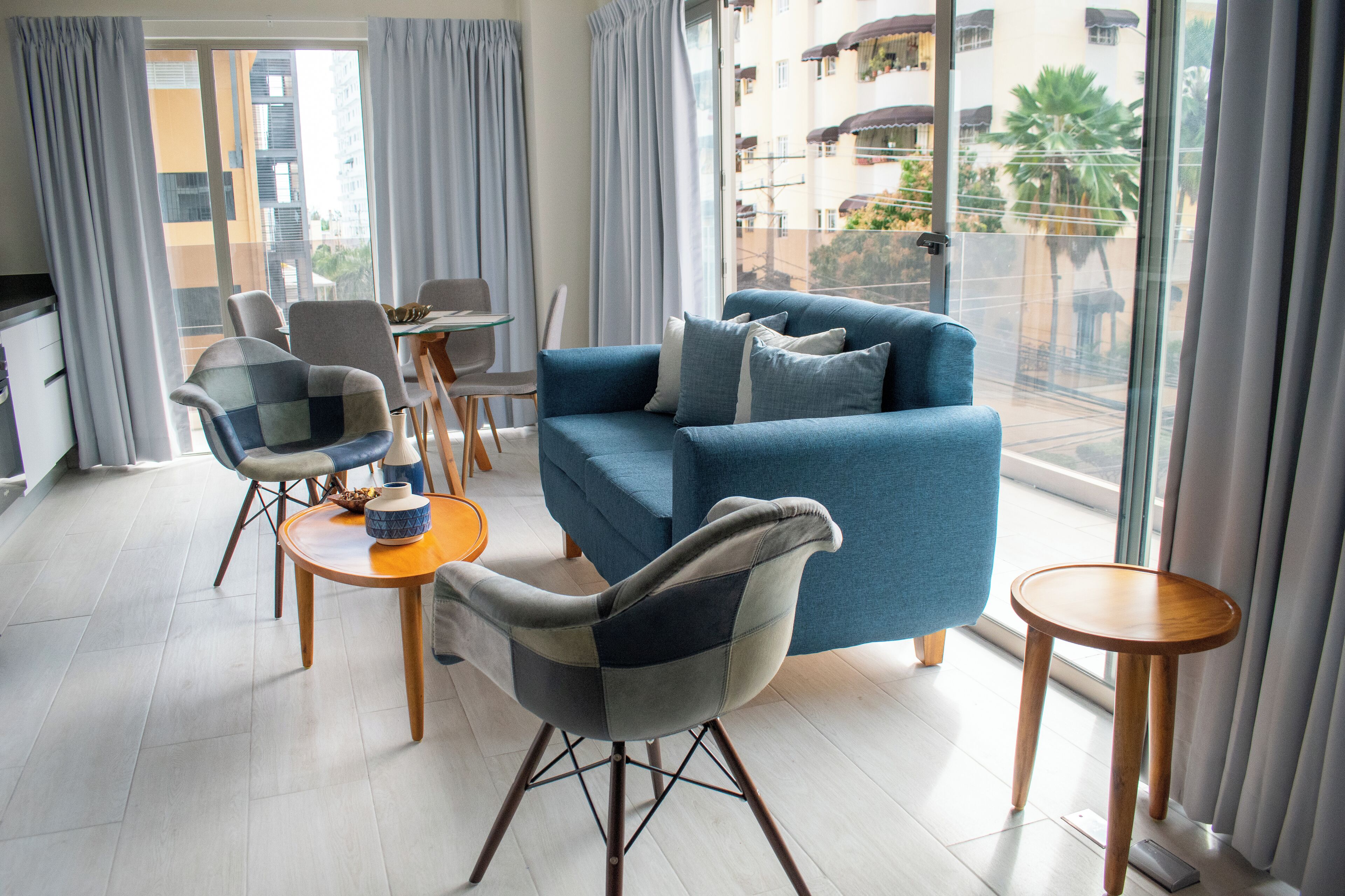 A modern living room with a blue sofa, two armchairs, a small table, and a view of a residential area through large windows.