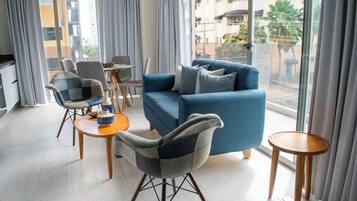 A modern living room with a blue sofa, two armchairs, a small table, and a view of a residential area through large windows.