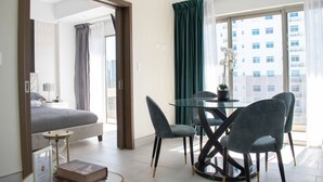 Moderni hotellihuone, jossa on sänky, lasinen ruokapöytä tuoleineen, pieni sivupöytä ja suuri ikkuna, josta avautuu näkymä kaupunkiin.