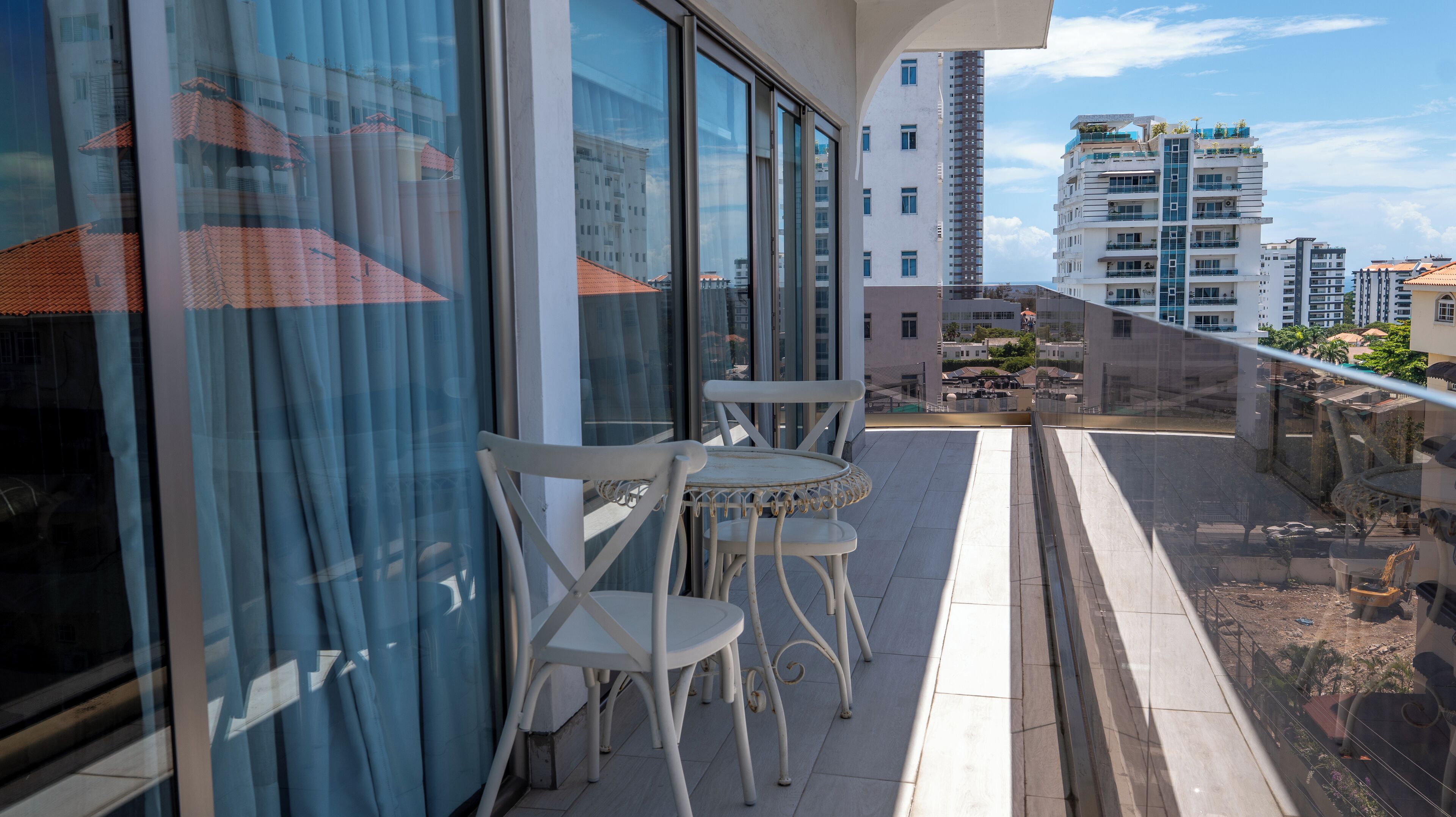 A balcony with two white chairs and a table, large windows, and a view of surrounding buildings.