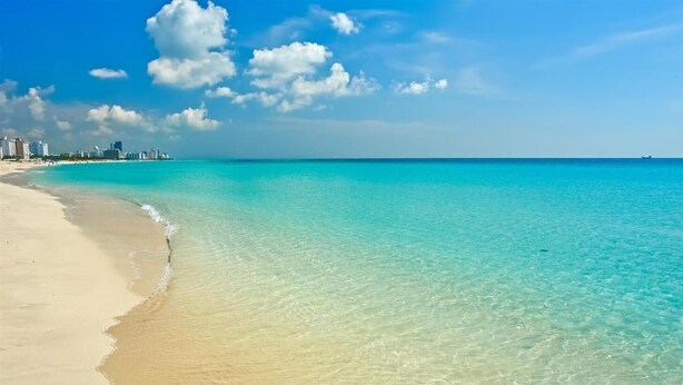 Een strand met helder turquoise water, een zandstrand en een stadsachtergrond.
