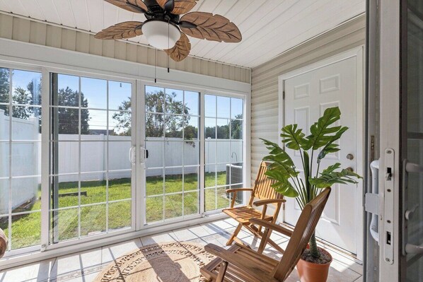 A sunroom with a sliding glass door, a ceiling fan, wooden chairs, and a potted plant.