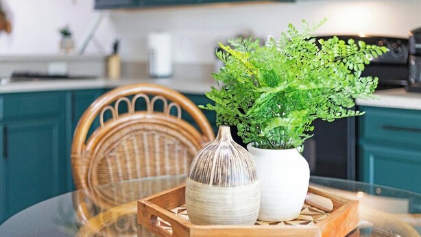 Un comedor con mesa de vidrio, una silla de mimbre, un florero blanco con follaje verde y una bandeja de madera.