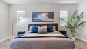 Un dormitorio con una cama, dos mesitas de noche, una lámpara de pie, un cuadro abstracto grande y una planta en maceta.