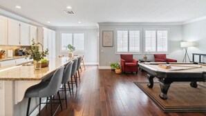 Amplia sala de estar con mesa de billar, comedor con taburetes de bar y cocina con alacenas blancas.