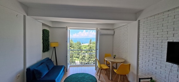 A room with a balcony, a sofa, a table with chairs, and a TV mounted on the wall.