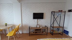 Une pièce avec un mur en briques blanches, un téléviseur à écran plat fixé au mur, un meuble télé en bois orné d’un cadre photo et surmonté d’une petite table, deux chaises jaunes et une petite table avec un pot de plante.
