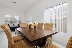 Un coin repas avec une table en bois et des chaises en osier, un vase rempli de fleurs et une vue par une fenêtre munie de stores.