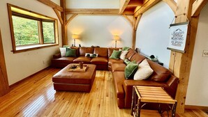 Ein gemütliches Wohnzimmer mit einer Eckcouch, Holzboden und Blick ins Freie durch große Fenster.