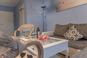 A living room with a white coffee table, a wicker chair with a 'home' pillow, a sofa with patterned cushions, and a wall-mounted artwork of birds. - 50010 Ferienhaus Eulennest - Das Ferienhaus "eulennest" mit 70 qm Wohnfläche Bietet Ausreichend (Wittmund)