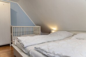 A bedroom with a white crib, a bed with white bedding, and a nightstand with a lamp. - 50010 Ferienhaus Eulennest - Das Ferienhaus "eulennest" mit 70 qm Wohnfläche Bietet Ausreichend (Wittmund)