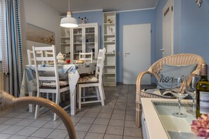 A dining area with a table set for two, a wicker chair with a pillow, and a wine bottle on a glass table.