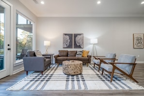 Amplia sala de estar con un gran ventanal, un sofá marrón, un sillón gris, una mesa de centro redonda y una alfombra estampada.