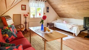 A cozy, well-lit attic bedroom with a wooden ceiling, a single bed, a small table with candles, a chair, and a colorful sofa.