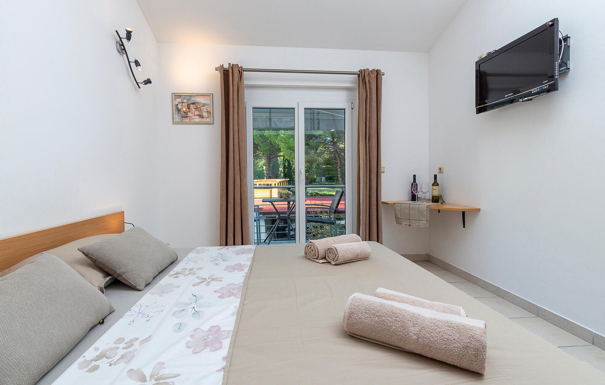 Une chambre moderne avec un lit, une télévision, un balcon avec une table et des chaises, et une vue sur la verdure.