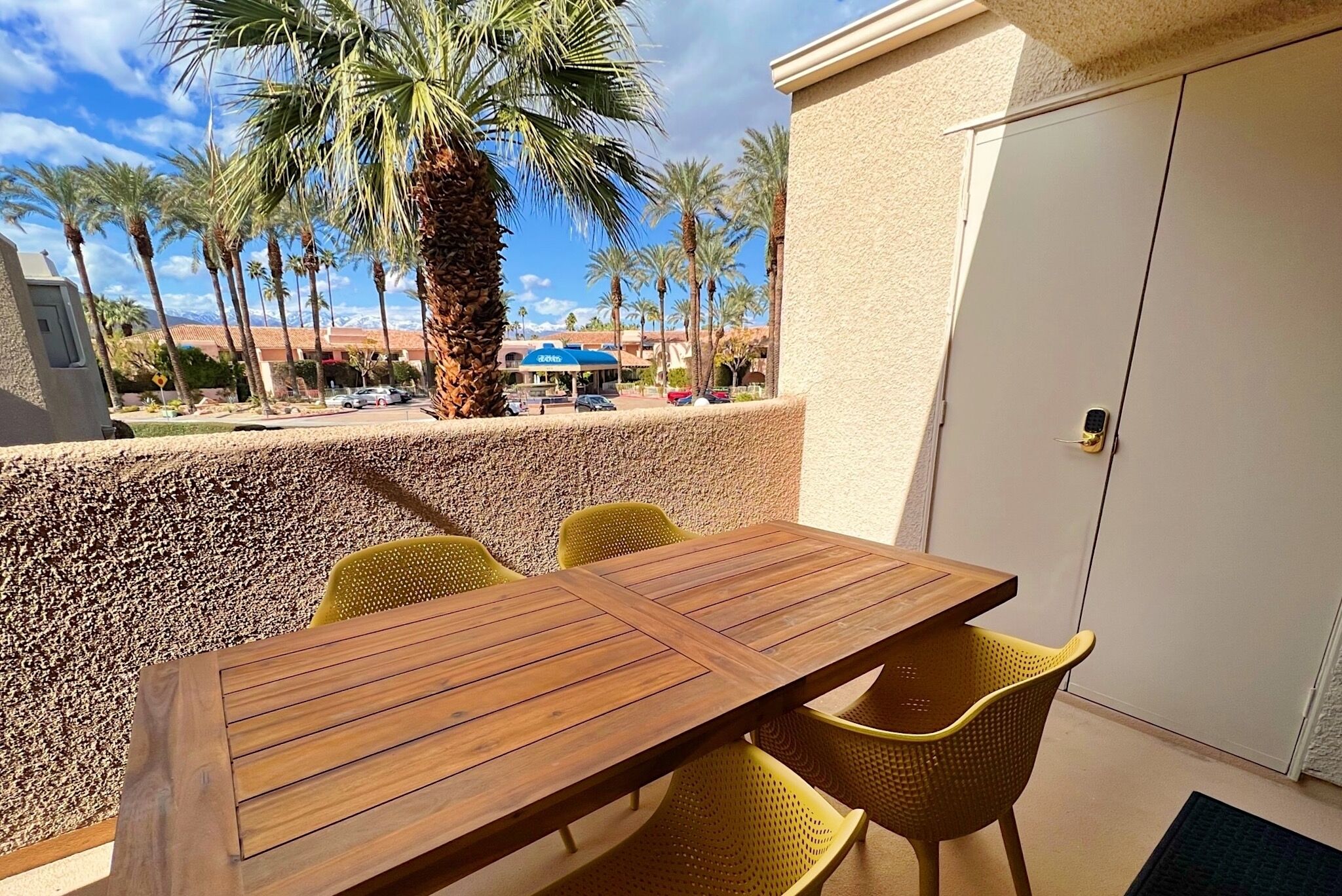 Une table en bois entourée de chaises jaunes sur une terrasse extérieure, avec en arrière-plan des palmiers et un bâtiment.