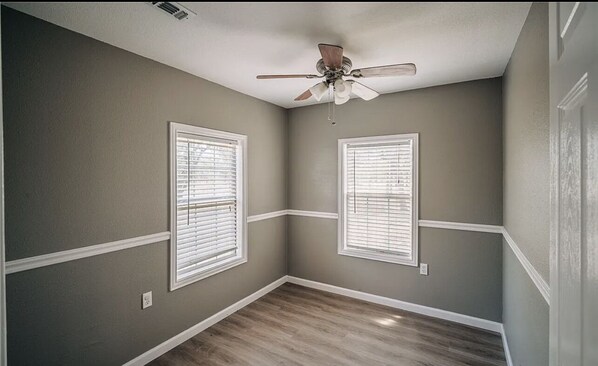 Una habitación con dos ventanas, ventilador de techo y suelo de madera.