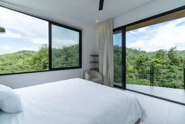 A bedroom with a large bed, a chair, and a view of a forested landscape through large windows.