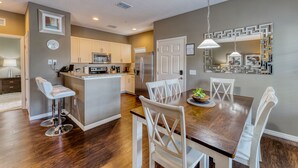 A modern kitchen with white cabinets, a central island, and a dining area with a wooden table and white chairs. - Beautiful Town Home in Gated Community With Amenities - Close to WDW (Kissimmee)