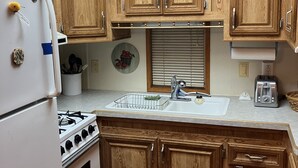 A compact kitchen with wooden cabinets, a white refrigerator, a gas stove, and a sink with a window above it. - Quaint and Quiet Tiny Home, with Pool Access! (Harlingen)