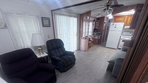Um espaço de estar compacto com sofá, poltrona e uma mesinha de centro com abajur. Uma cozinha é visível ao fundo, com eletrodomésticos e uma mesa de jantar.