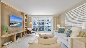A modern hotel room with a large window, a flat-screen TV, a sofa, a chair, and a bookshelf.