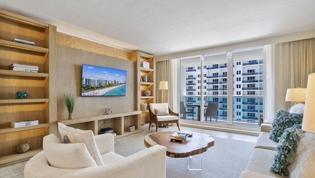 A modern hotel living room with a large TV, comfortable seating, a wooden coffee table, and a view of a cityscape through large windows.
