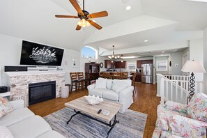 A spacious living room with a fireplace, a television, a ceiling fan, and a dining area with a kitchen in the background.