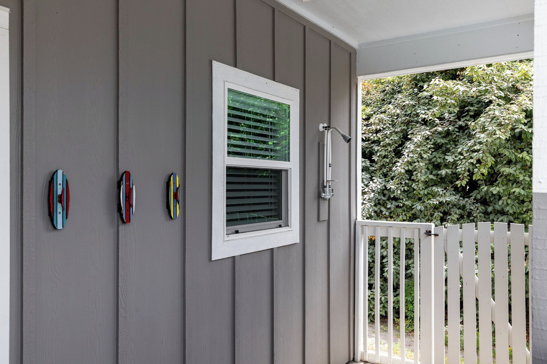 Pared exterior gris con una ventana blanca, una cerca de piquetes blancos y tres objetos coloridos sujetos a la pared.
