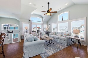 A spacious living room with a ceiling fan, large windows, and a view of the outside. - TJs Beach Bungalow:Private & community pool, hot tub, dog friendly and elevator! (Corolla)