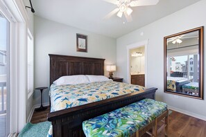 A bedroom with a wooden bed, a bench, a ceiling fan, a mirror, and a view of the outside.