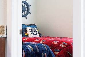A bedroom with a bed featuring a nautical-themed duvet and pillow, a wall-mounted ship's wheel decoration, and a partial view of a wooden door. - TJs Beach Bungalow:Private & community pool, hot tub, dog friendly and elevator! (Corolla)