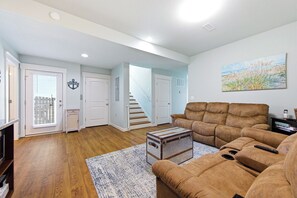 A living room with a brown sofa, a wooden floor, a white door, and a staircase leading upstairs. - TJs Beach Bungalow:Private & community pool, hot tub, dog friendly and elevator! (Corolla)
