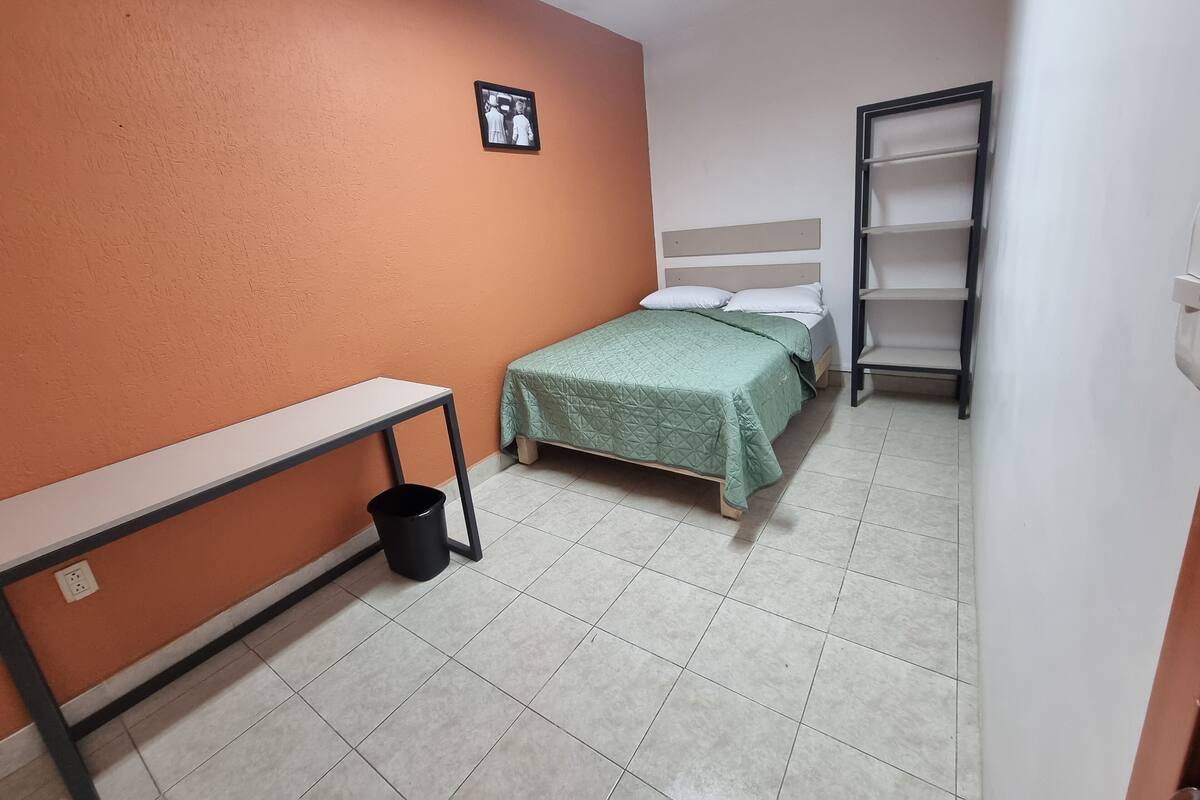 Quarto pequeno e bem arrumado, com uma cama de solteiro, mesa de cabeceira, lixeira e prateleira na parede.
