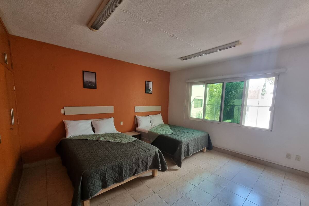 Quarto com duas camas, janela com vista para o verde e teto branco com ventilação.