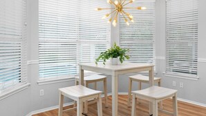 A dining area with a white table and four chairs, surrounded by large windows with blinds. - Incredible Heated Pool/HotTub! Gameroom-Movieroom! (Pflugerville)