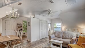 Un salon spacieux avec un plafond haut, un lustre, un ventilateur de plafond, un coin repas avec une table en bois et des chaises blanches, et un canapé beige.