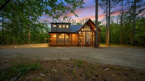 A two-story log cabin with a stone chimney, surrounded by a gravel driveway and forest. - Game Room, Hot Tub, Fire Pit, Pets, High Horse (Broken Bow)
