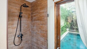 Ducha con cabezal de ducha de mano y manguera, junto a una puerta de cristal que da a una piscina.