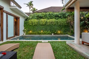 Una piscina con un patio circundante, una pared de vegetación y un edificio con techo de tejas.
