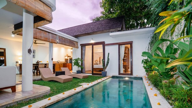 Una piscina con un borde perimetral, un área de comedor con sillas y mesas, y vistas del interior a través de puertas de cristal.